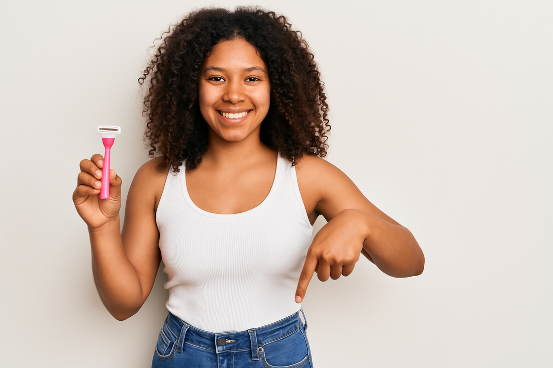 Natürlich aussehendes Black Girl mit strahlender Haut, lockigen Haaren und weißem Top hält einen pinken Rasierer in der Hand und zeigt mit dem Finger auf ihren Intimbereich, trägt eine blaue Jeans, heller Studiohintergrund.
