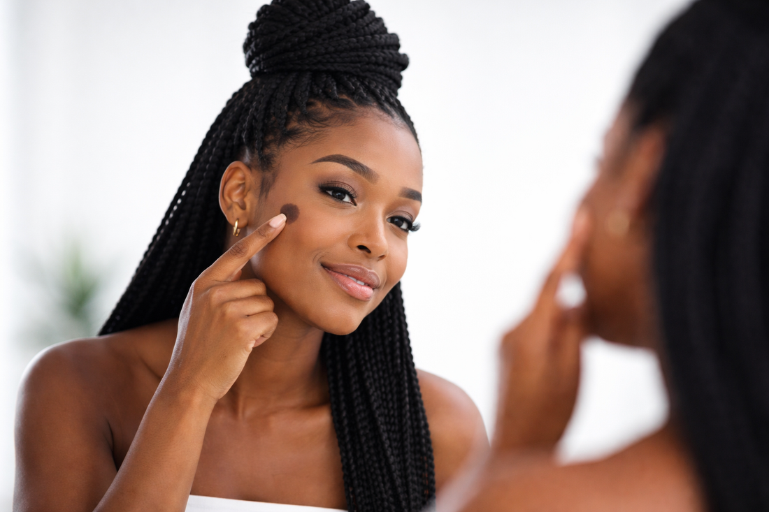 Black-Skin-Frau mit Braids betrachtet einen Pigmentfleck auf der Wange im Spiegel vor weiß-grauem Hintergrund