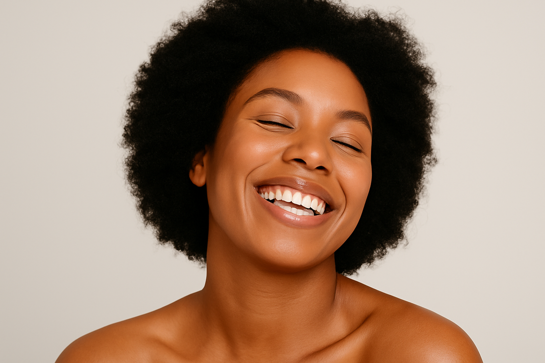 Dunkelhäutige Frau mit natürlichem Afro lächelt breit vor hellem Hintergrund; warmer, weicher Beauty-Portrait-Look im Querformat.
